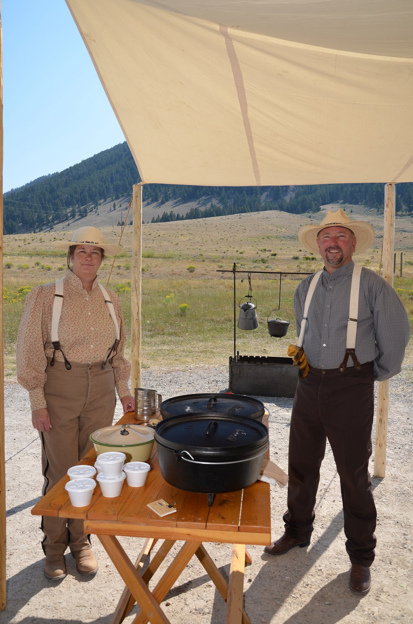 Two men in period range dress standing at a wooden table under a canvas fly, a cast iron dutch oven and stacked enamel cups on the table in front of them, open country and hills behind.