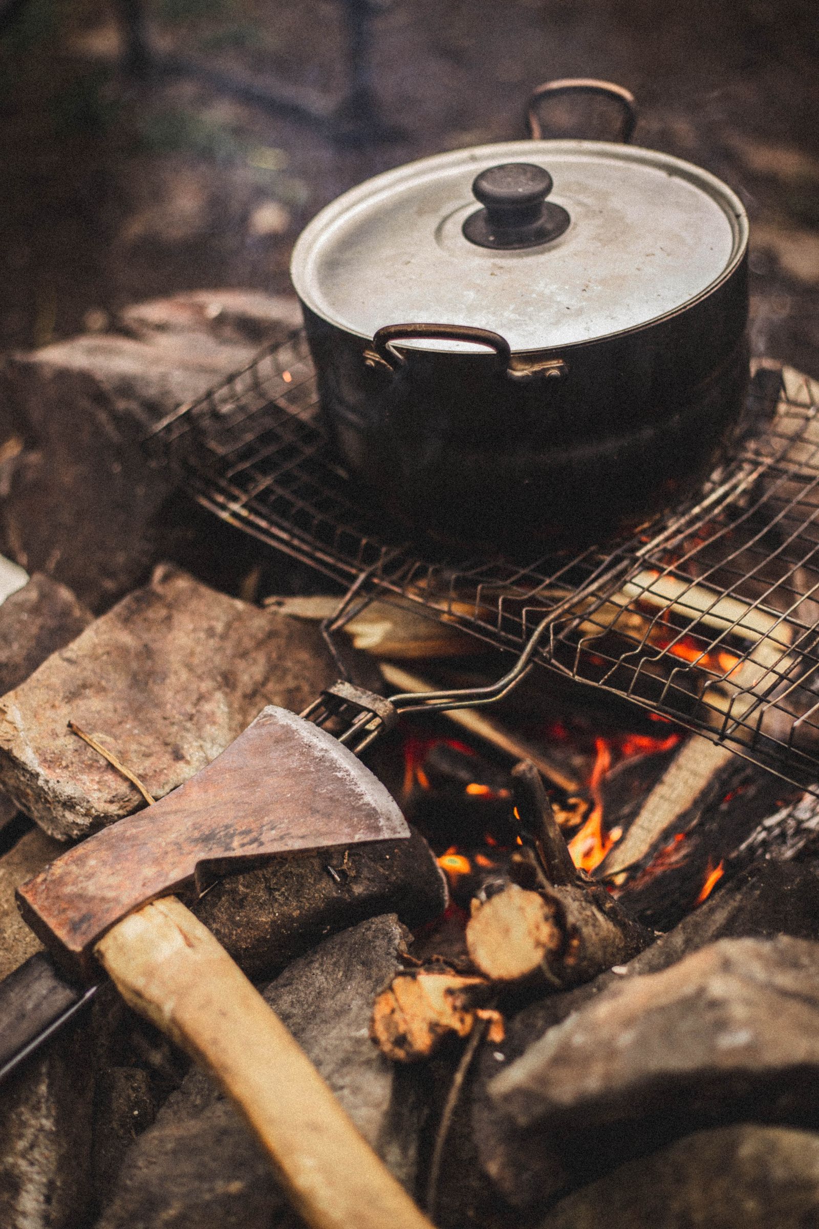 Cast iron pot over a campfire with a hatchet resting against a log in the foreground, camp cooking in an outdoor setting.