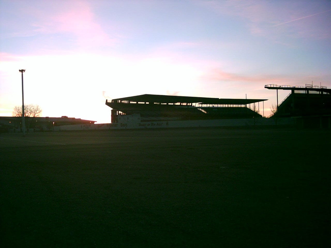 The Frontier Park grandstand in silhouette at dusk, with the empty arena infield in the foreground and a pink and grey Wyoming sky behind.