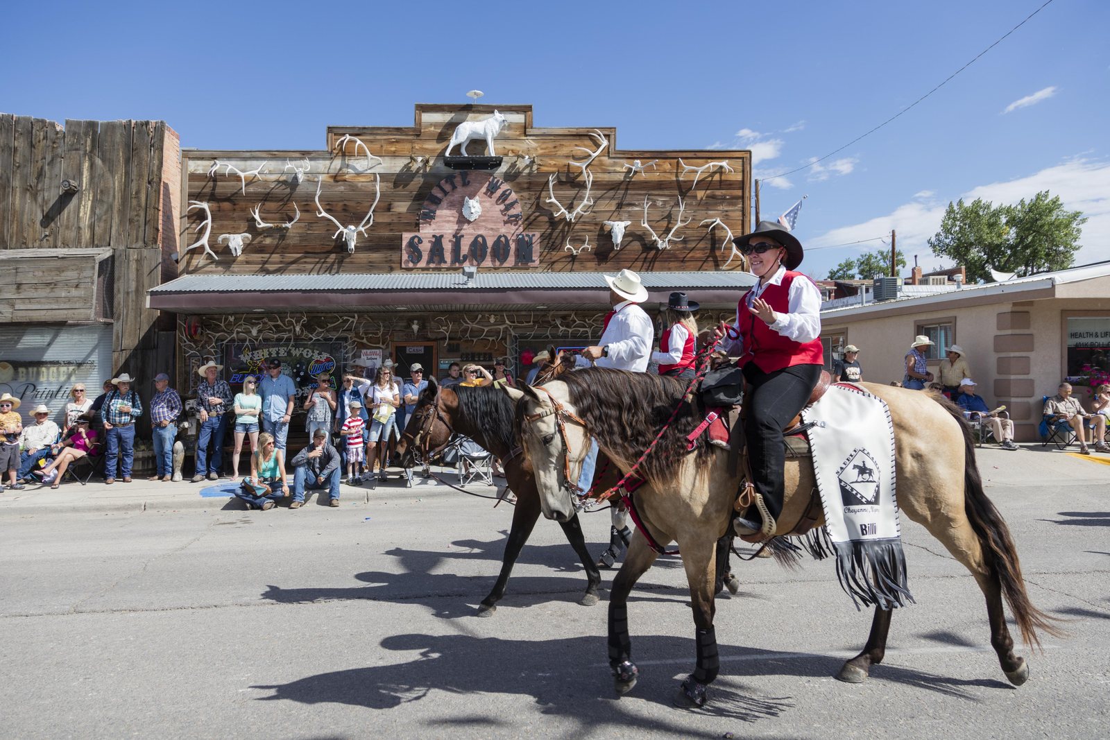 Two riders in sashes and white hats on horseback in the middle of a Douglas, Wyoming street during the state fair parade, passing a crowd seated along the curb outside a saloon front hung with antlers.