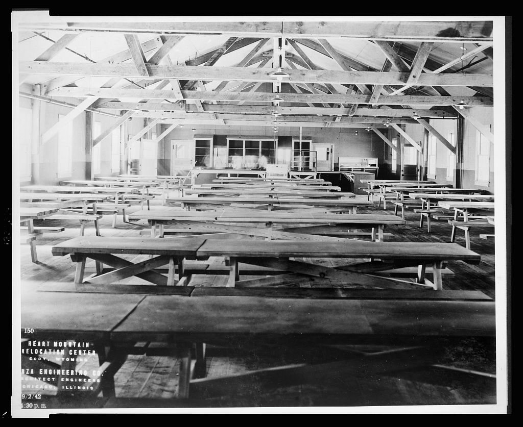 A 1942 black and white photograph of a newly built mess hall interior at Heart Mountain Relocation Center, rows of long bench tables under exposed roof trusses and bare bulbs.