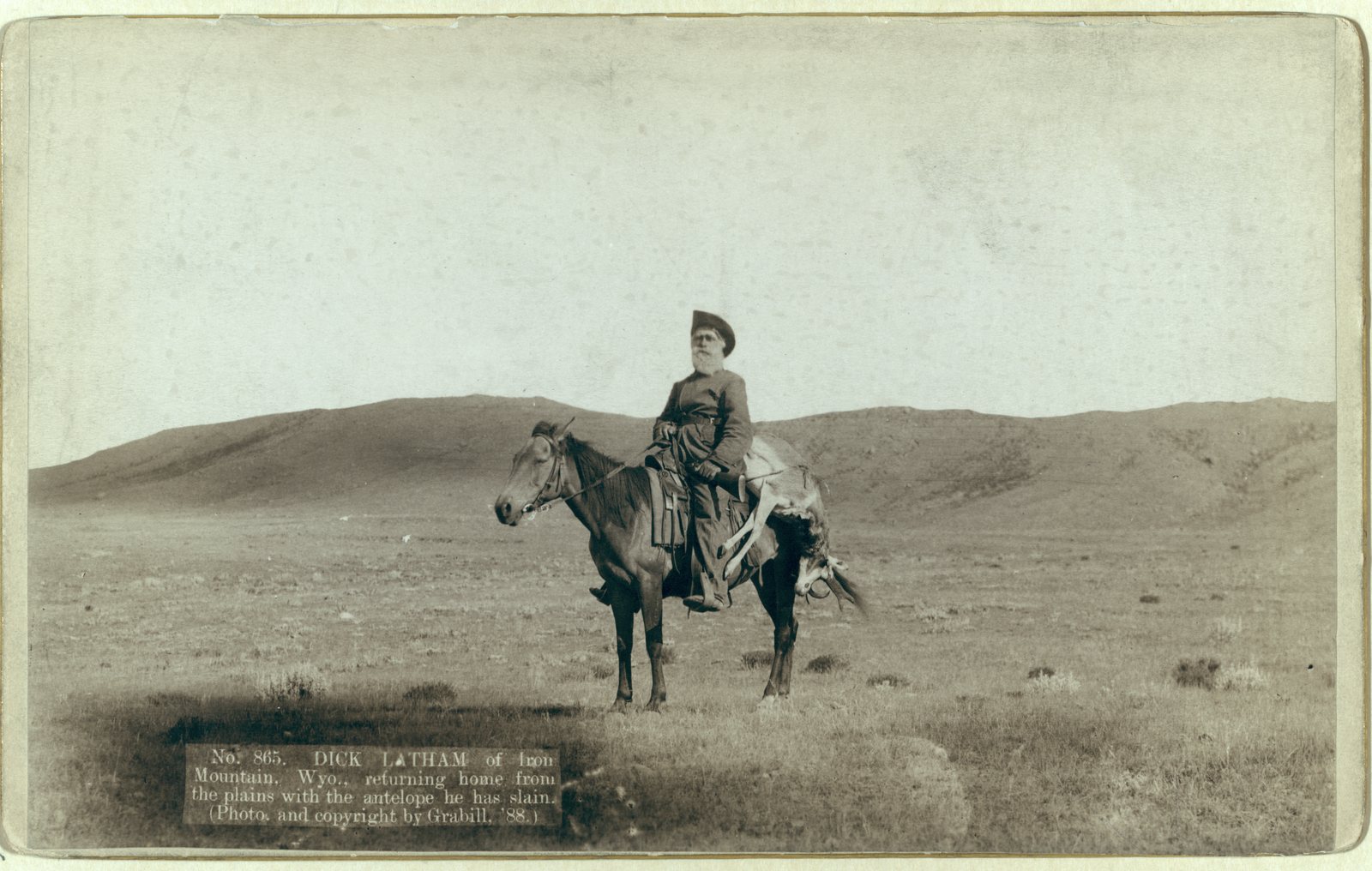 Cabinet-card photograph of a lone rider crossing bare sagebrush plains with a dead antelope slung behind his saddle, a long low ridge running across the horizon.