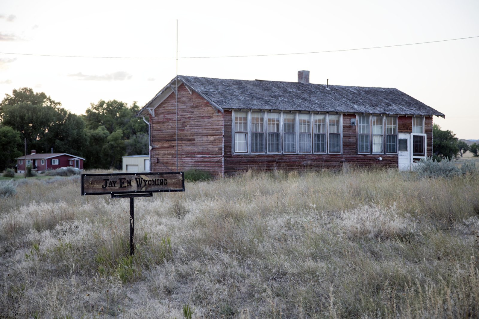 A long weathered wooden building with boarded and broken windows standing behind waist-high dry grass at Jay Em, Wyoming, with a small hand-lettered sign in front of it.