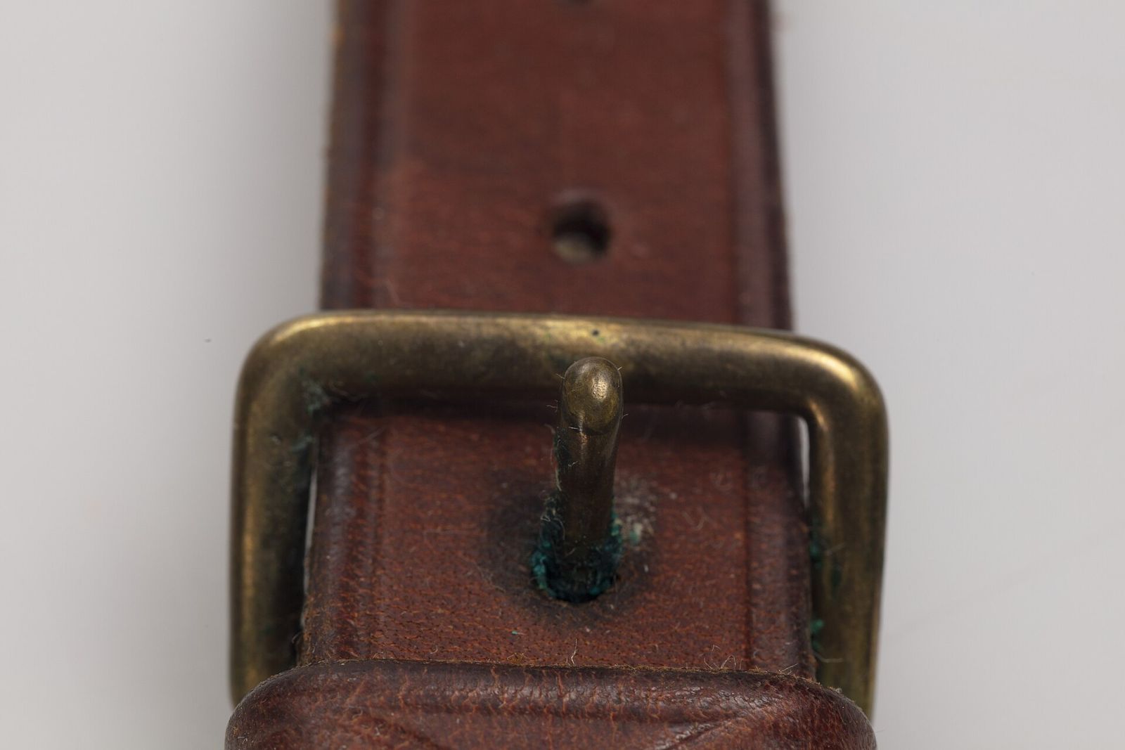 A plain brass buckle on a brown leather belt, with a hand-stitched keeper and a small green spot of verdigris near the prong.