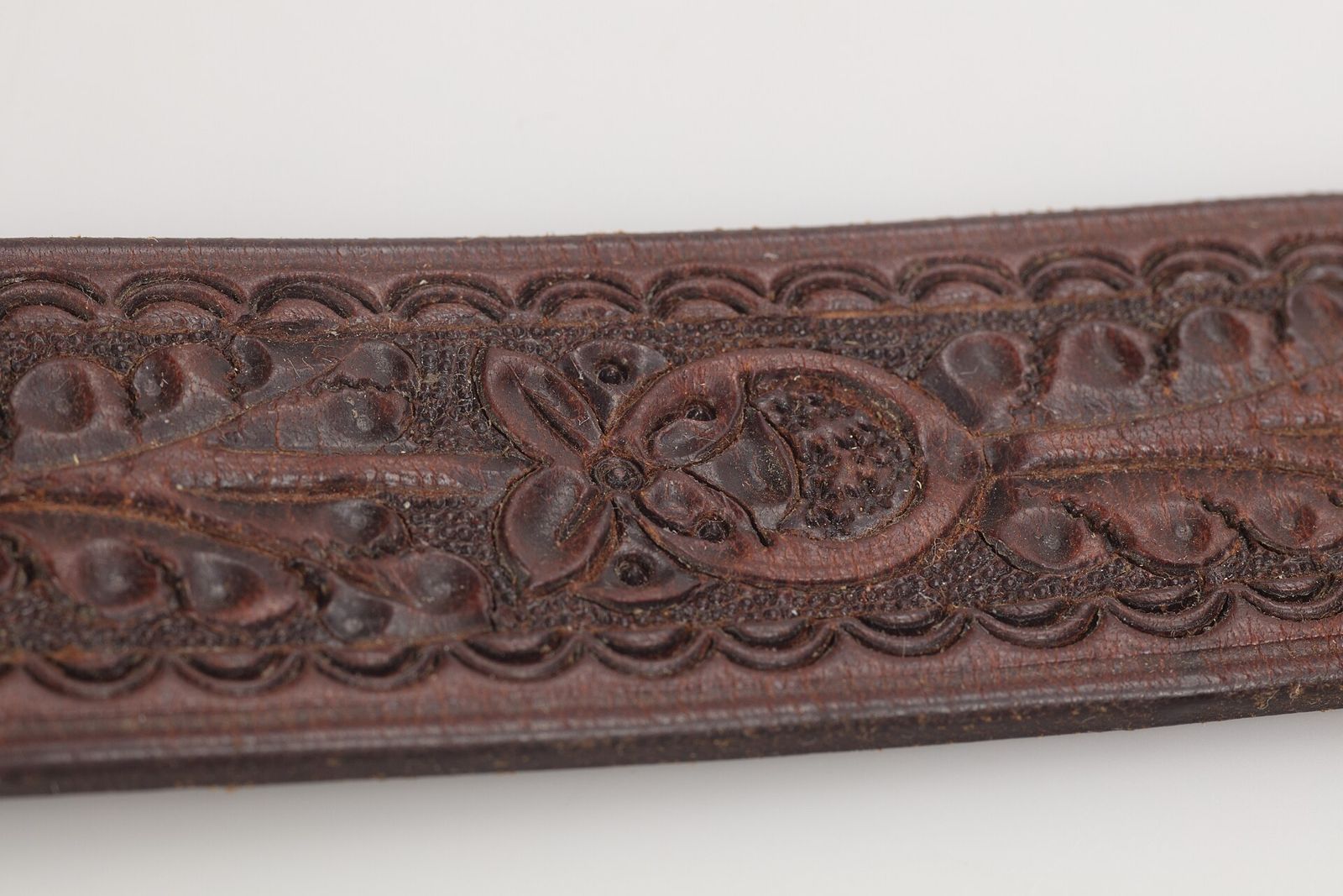 Close view of a dark brown leather belt with deeply carved floral and scroll designs between two bands of repeated border stamping.