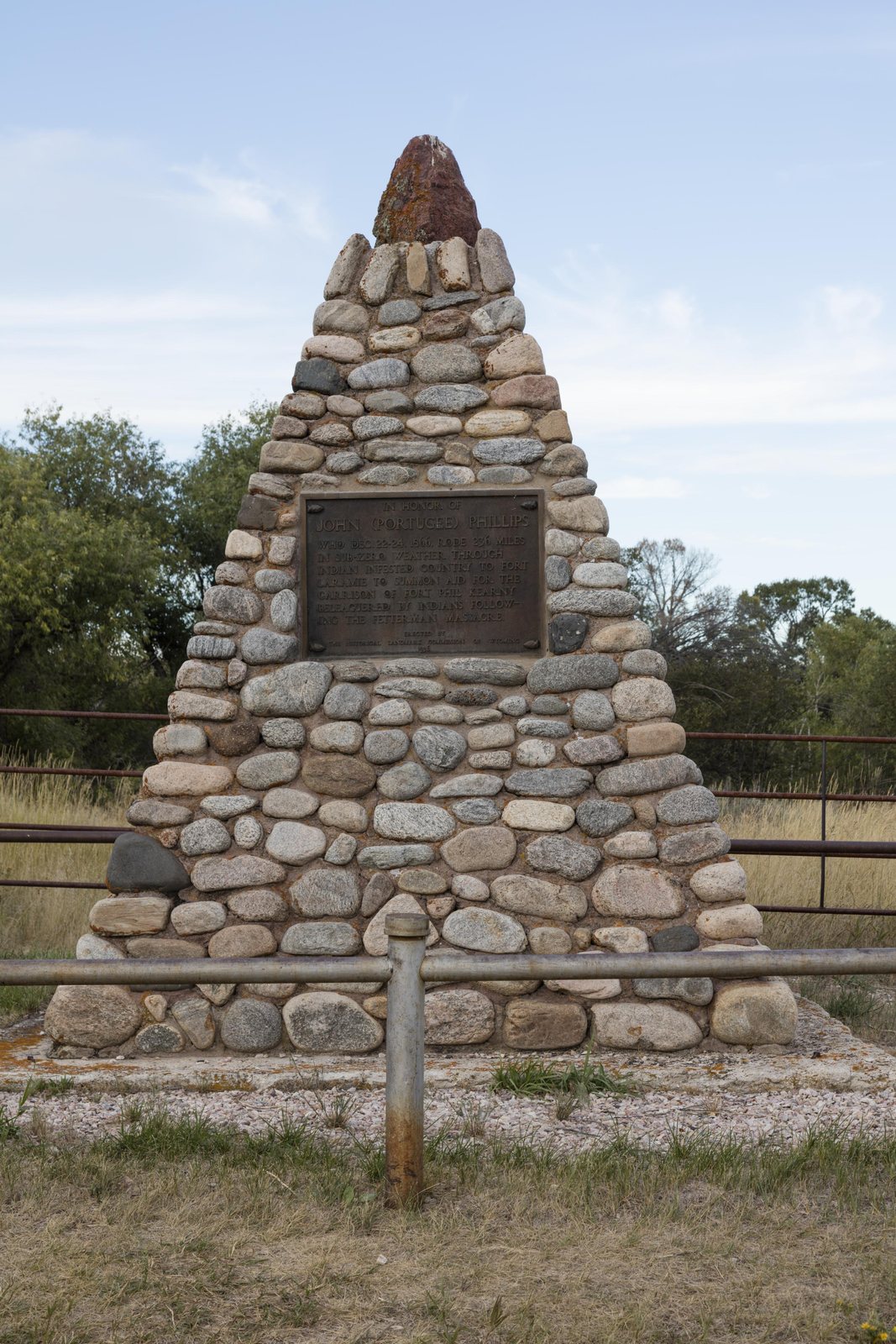 Tall pyramid of rounded river cobbles mortared together, a dark bronze plaque set into the front face, standing in mown grass behind a pipe rail.