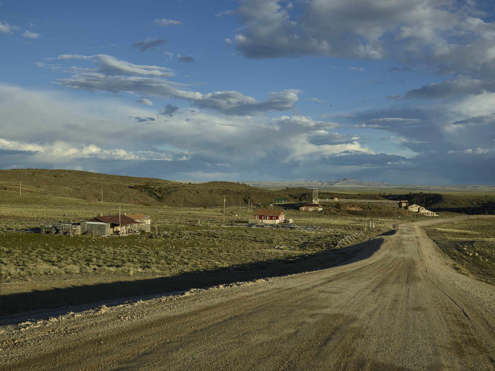 A gravel road running across open sage country toward the scattered low buildings of South Pass City, Wyoming, under a wide sky of broken cloud.