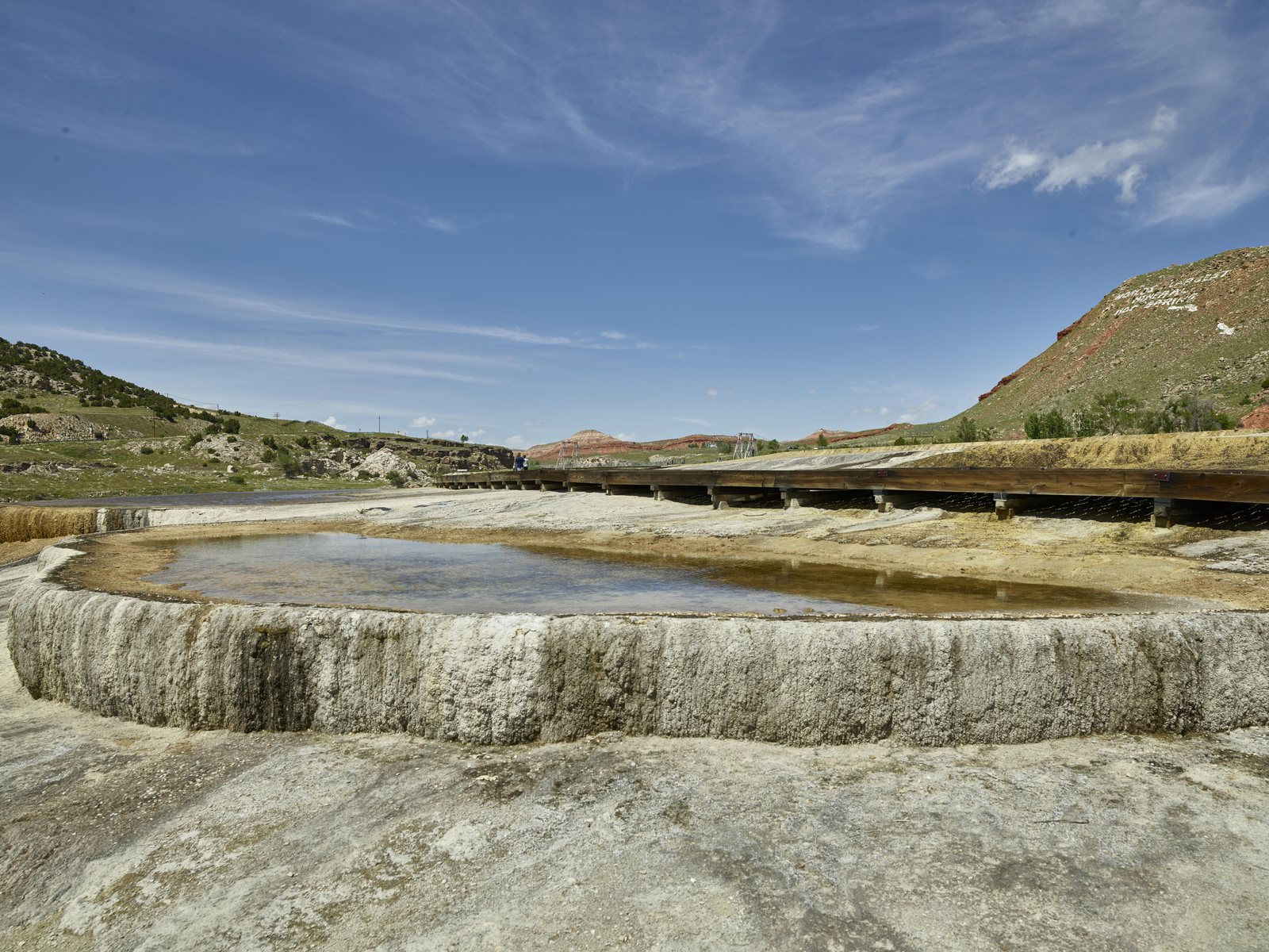 A wide travertine terrace holding a still green pool at Hot Springs State Park in Thermopolis, Wyoming, with a footbridge and low red hills behind it.