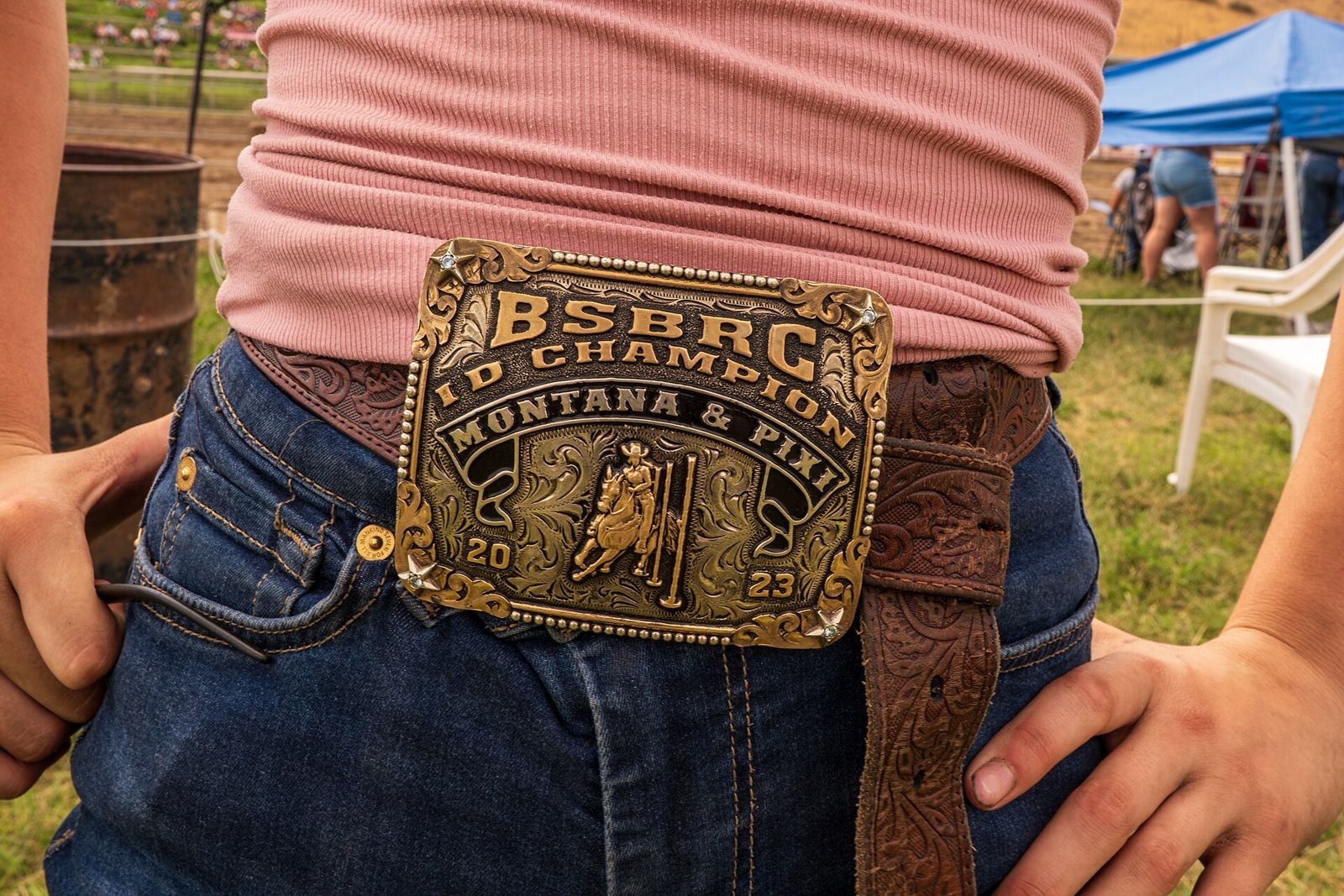 A youth rodeo champion's engraved trophy buckle, reading Montana and Pixi 2023, worn on a tooled brown leather belt with blue jeans.