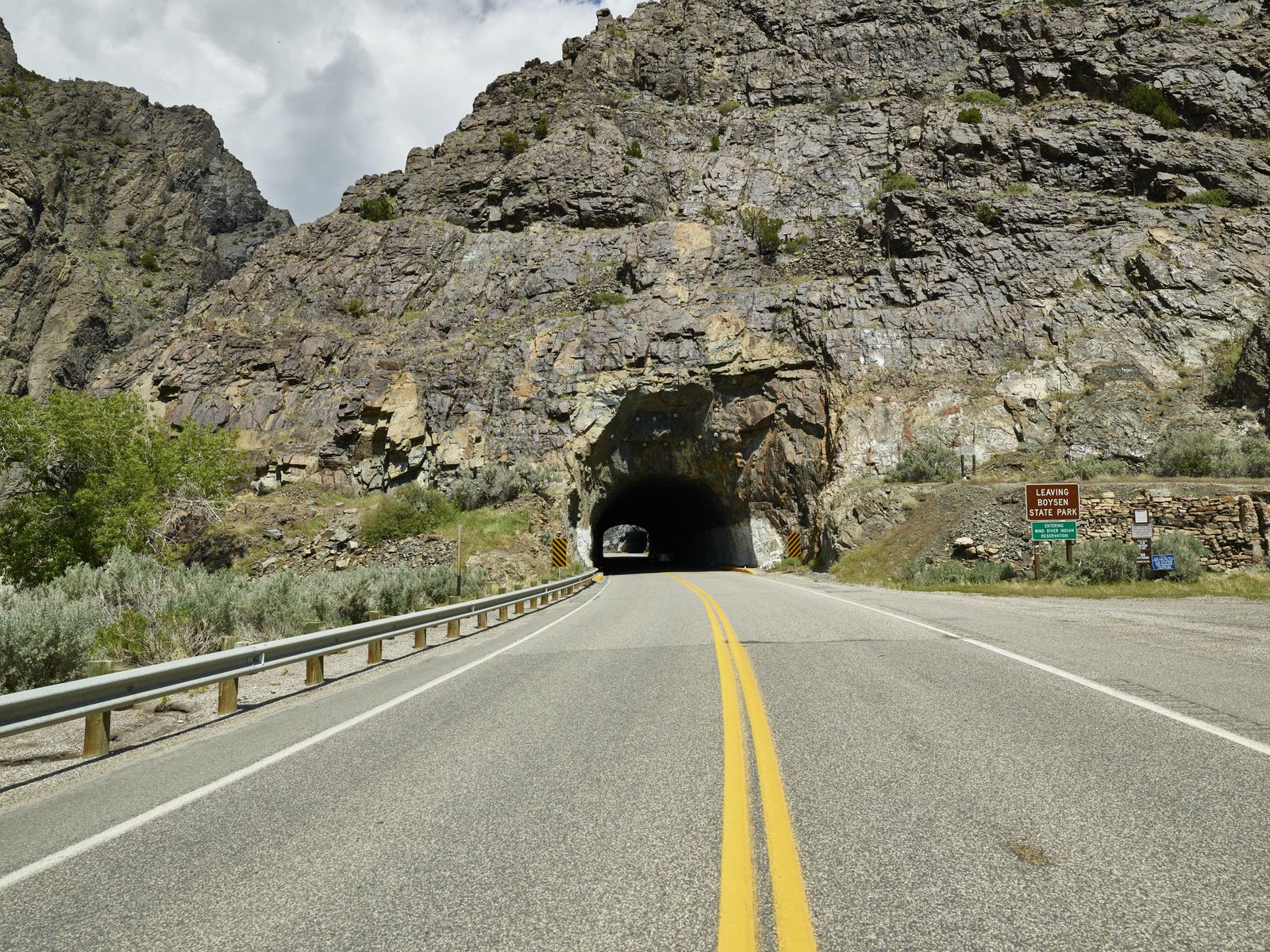 A two-lane highway with a double yellow center line running along a guardrail toward a rough-cut rock tunnel in the wall of the Wind River Canyon, Wyoming.