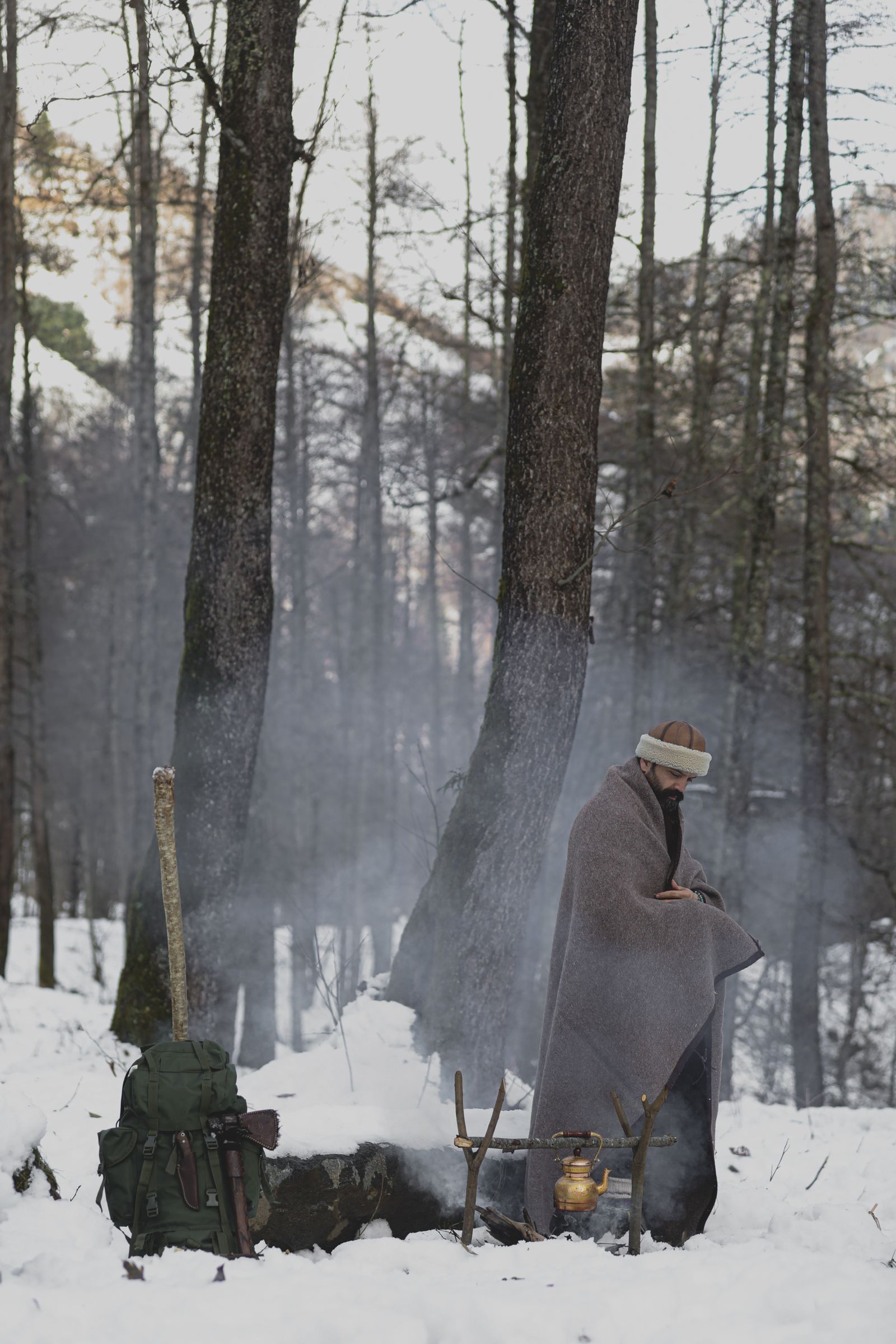 A person wrapped in a wool blanket sitting by a campfire in an outdoor setting, the classic working context for a heavyweight wool camp blanket.