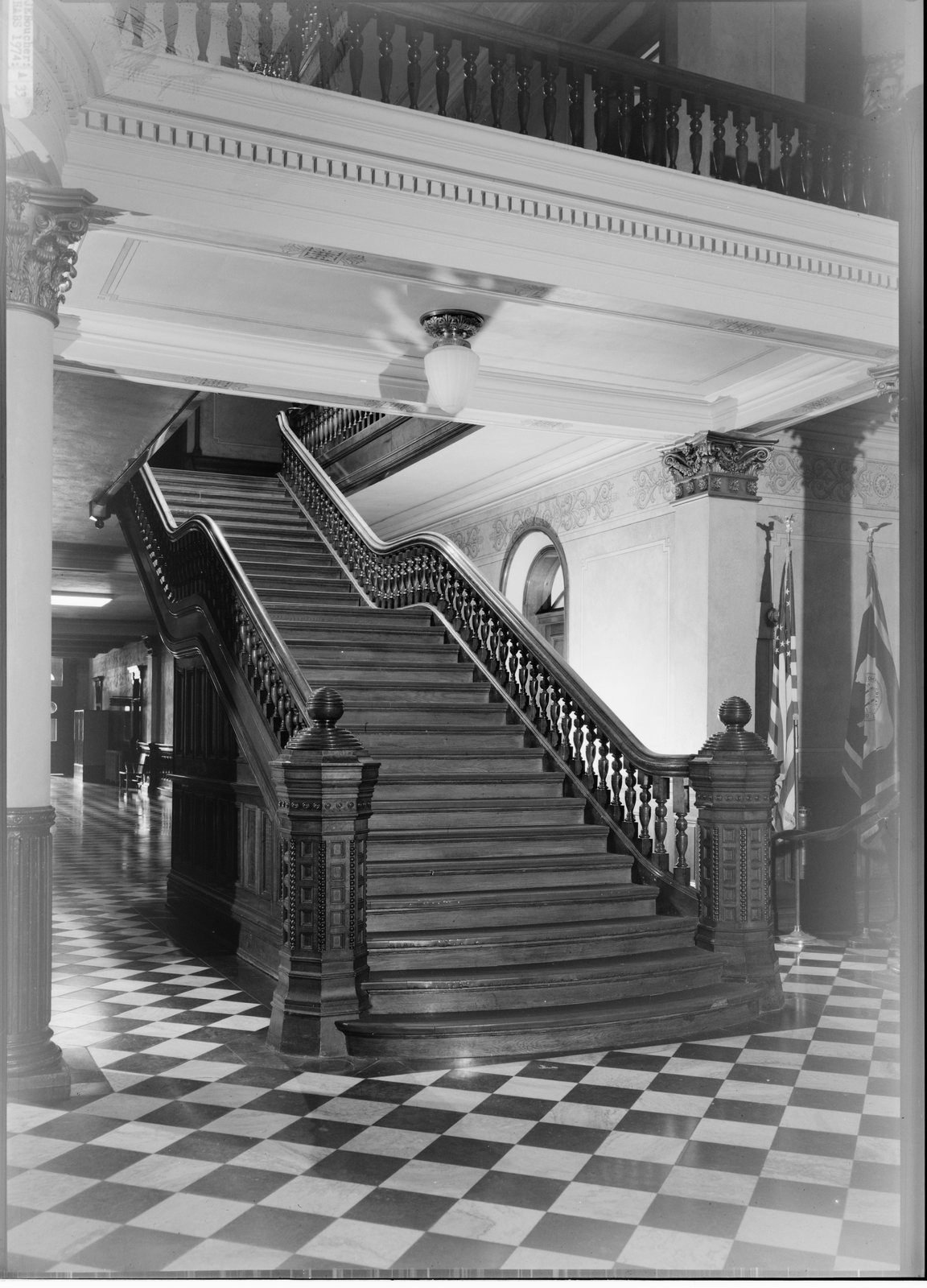 Wide dark wooden staircase with heavy carved newel posts and turned balusters rising from a black-and-white checkerboard marble floor, flags and a fluted column at the right.