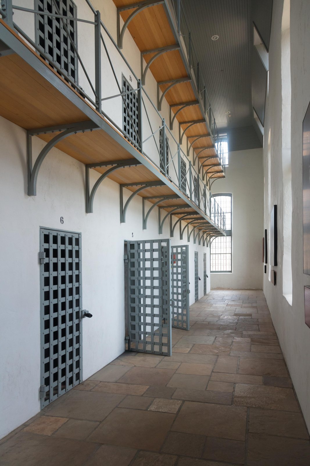Long narrow cellblock corridor with a tiled floor, a row of open latticed steel cell doors on the left, and a second tier of cells on a railed gallery above.