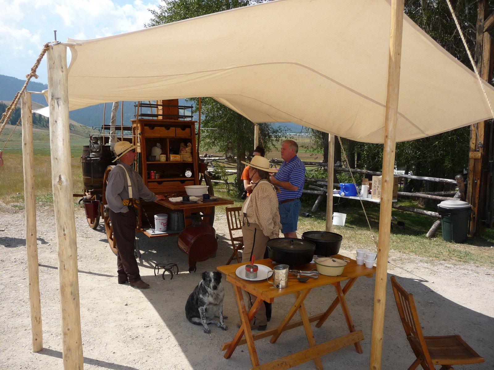 Two cooks working under a canvas fly at a wooden chuckwagon, cast-iron Dutch ovens and enamel plates set out on a folding table, a dog lying underneath in the shade.