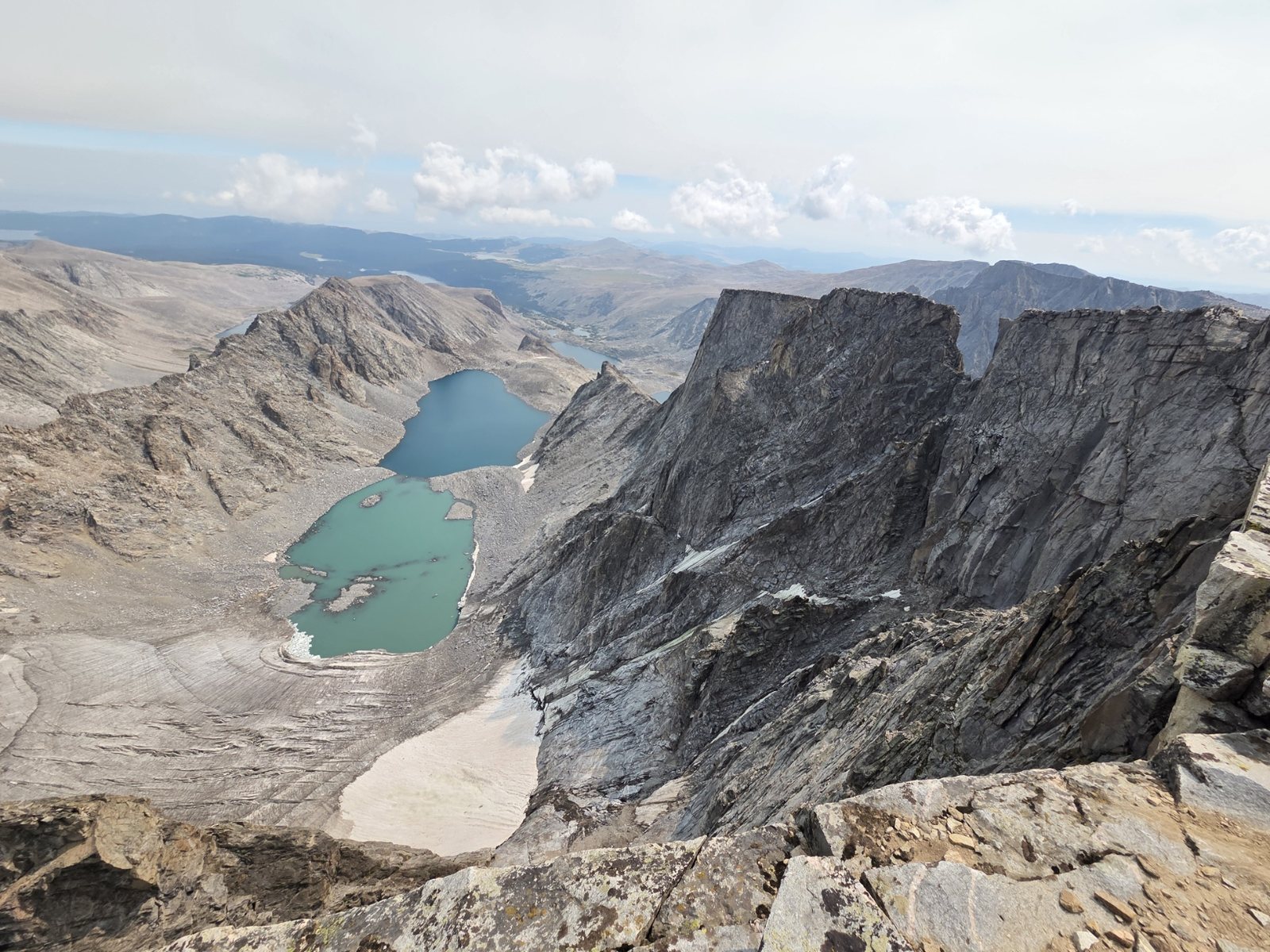 The view east from the summit of Cloud Peak in the Bighorn Mountains, Wyoming, looking down a shattered rock wall onto two green alpine lakes with a snowfield beside them.