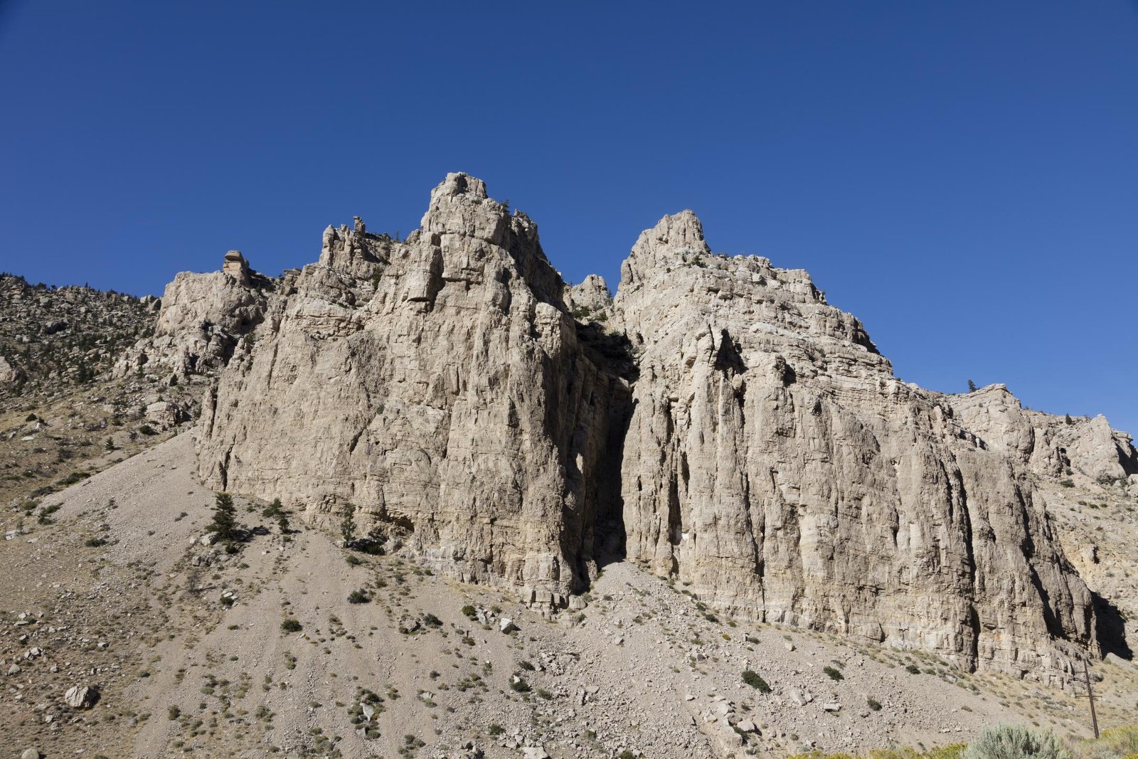 Pale limestone cliffs rising in blocky towers above a scree slope of sagebrush and loose rock in Shoshone Canyon, Park County, Wyoming, under a hard blue sky.