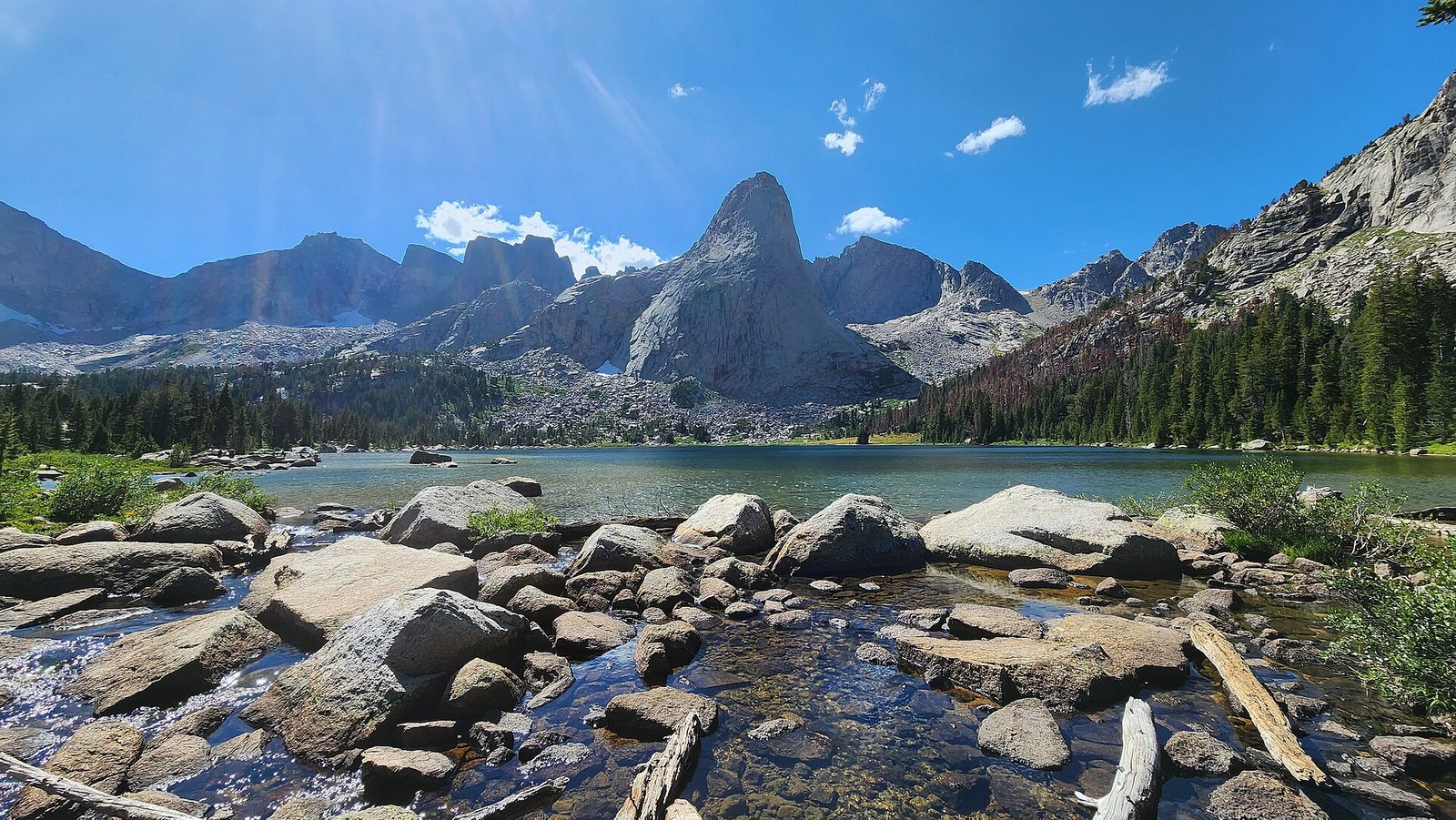 The granite spires of the Cirque of the Towers rising above Lonesome Lake, with a boulder-choked outlet stream in the foreground and conifers along the shore.