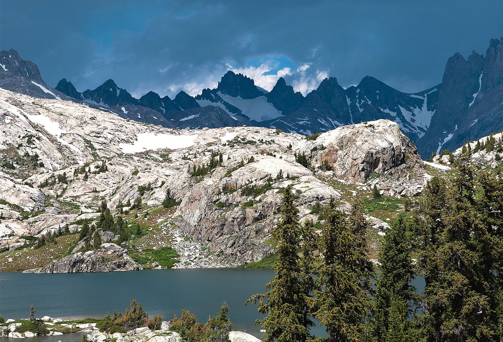 A storm building over granite walls and snowfields above a lake in Titcomb Basin, with polished slab and stunted conifers in the foreground.